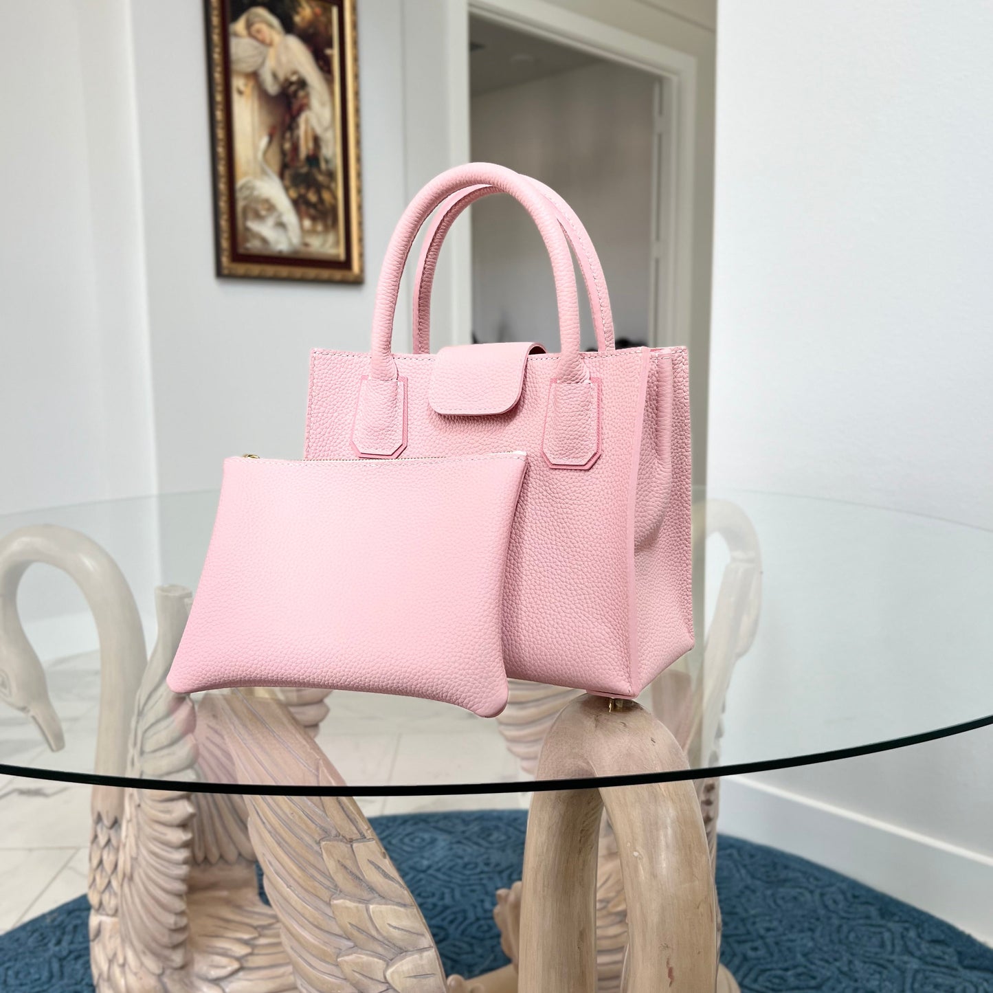 Pink handbag and pouch on a glass table with a white wall and painting in the background